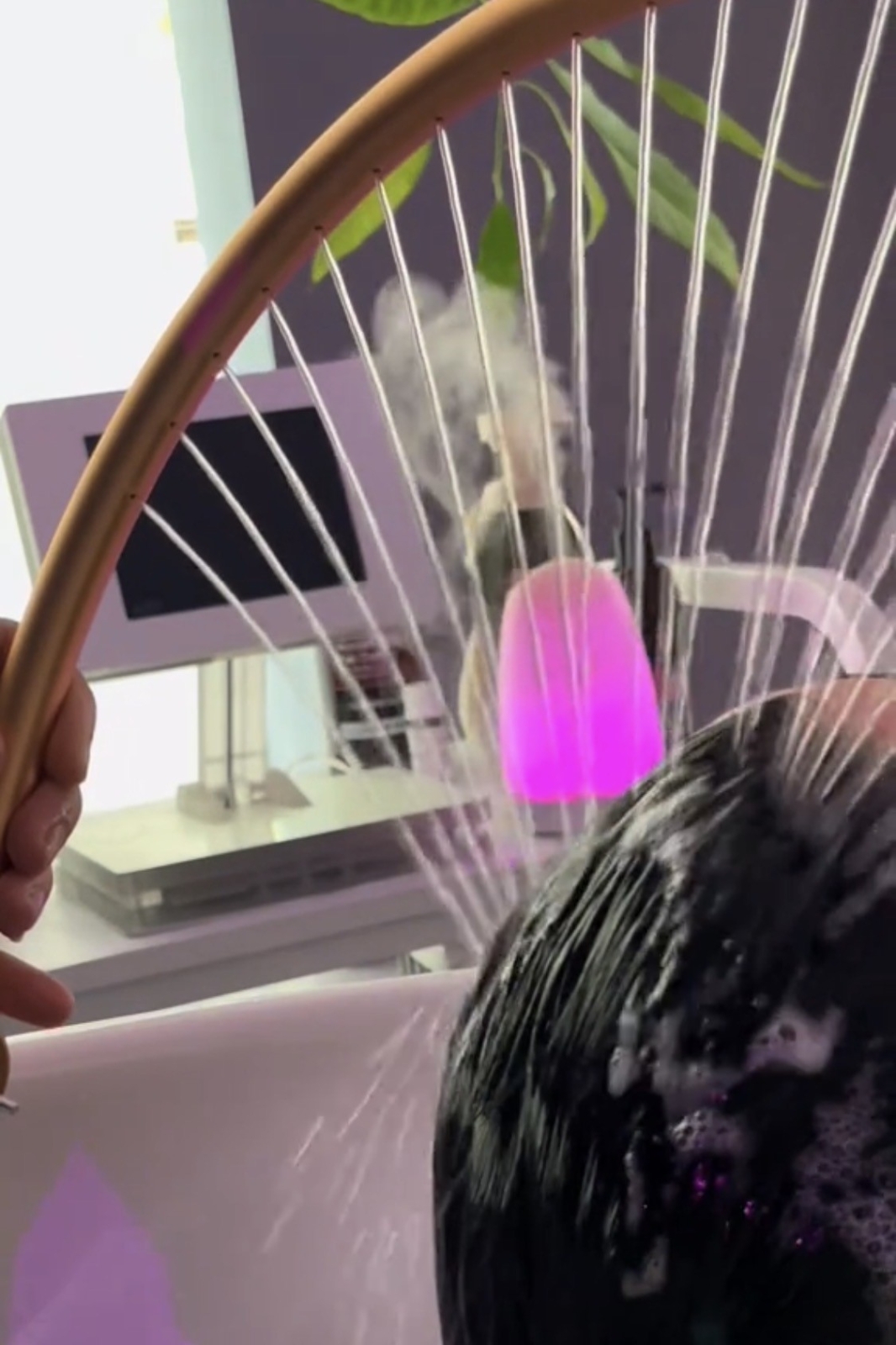 Close-up of a curved wooden hoop with many thin metal rods radiating outward, pink light in the background, with wet dark hair in the foreground.