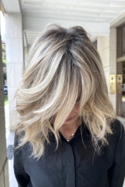 IMG_8338 Close-up of a person with long, wavy blonde hair partially covering their face, wearing a black shirt outdoors beside a light building.