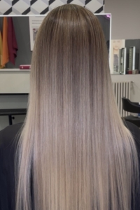 Back view of a person with long, straight hair transitioning from dark at the roots to platinum blonde at the ends, in a hair salon.