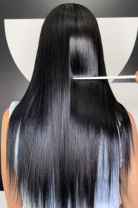 Back view of a person with long, glossy black hair being cut by a stylist using a white comb and scissors in a studio setting.