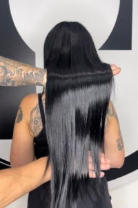 Person with long, straight black hair held down to show length; visible arm tattoos and black top in a salon setting.