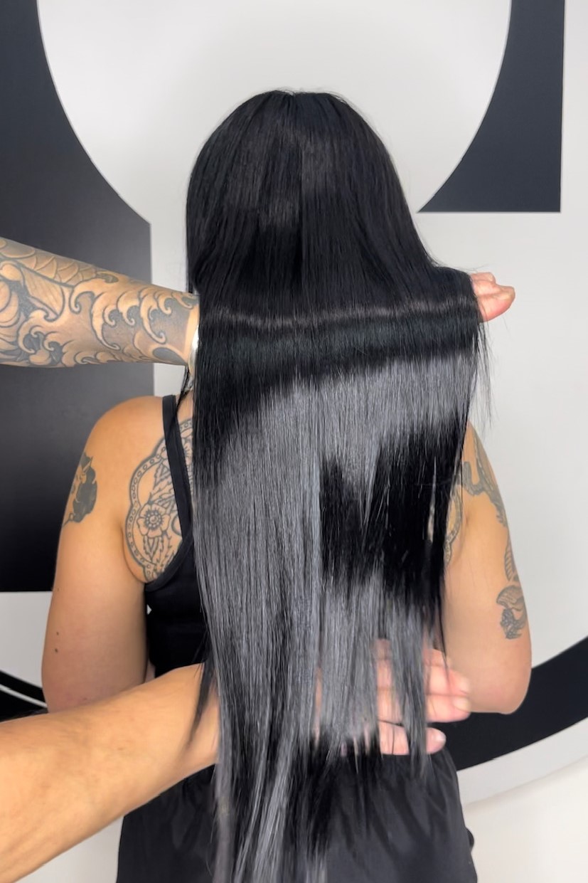 Person with long, straight black hair held down to show length; visible arm tattoos and black top in a salon setting.