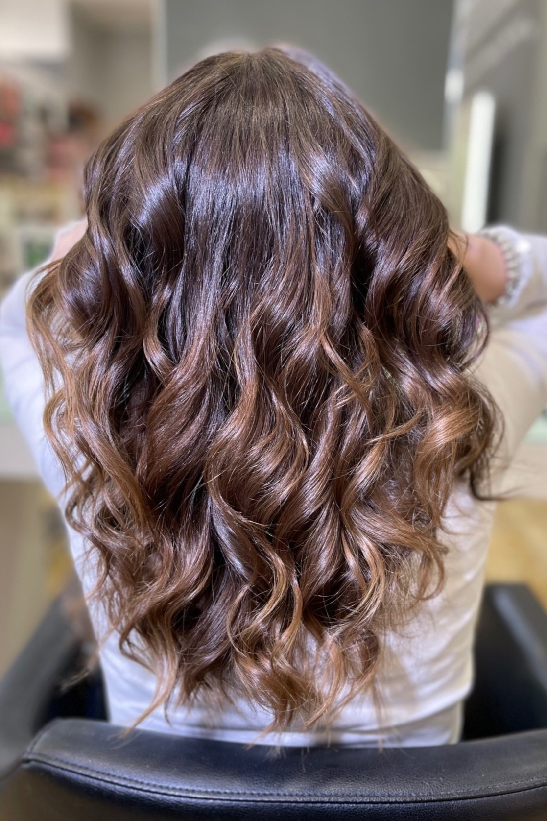 Back view of a person with shoulder-length wavy brown hair styled in loose curls at a salon. The hair looks glossy with subtle highlights in a professional setting.