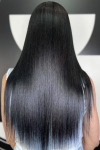 Back view of a person with long, straight, glossy black hair reaching mid-back, wearing a light denim top against a neutral background.
