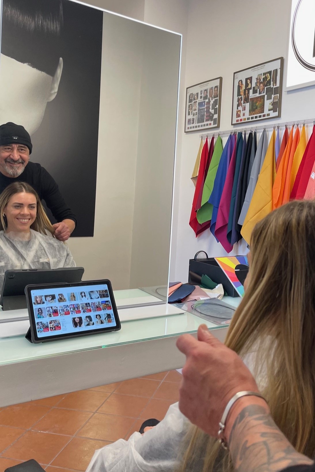 Hairdresser standing behind a smiling client seated in a salon chair, tablet showing a photo gallery on the counter in front of them.