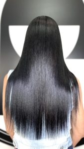 Back view of a person with long, sleek black hair reaching mid-back, wearing a light denim jacket against a dark background.